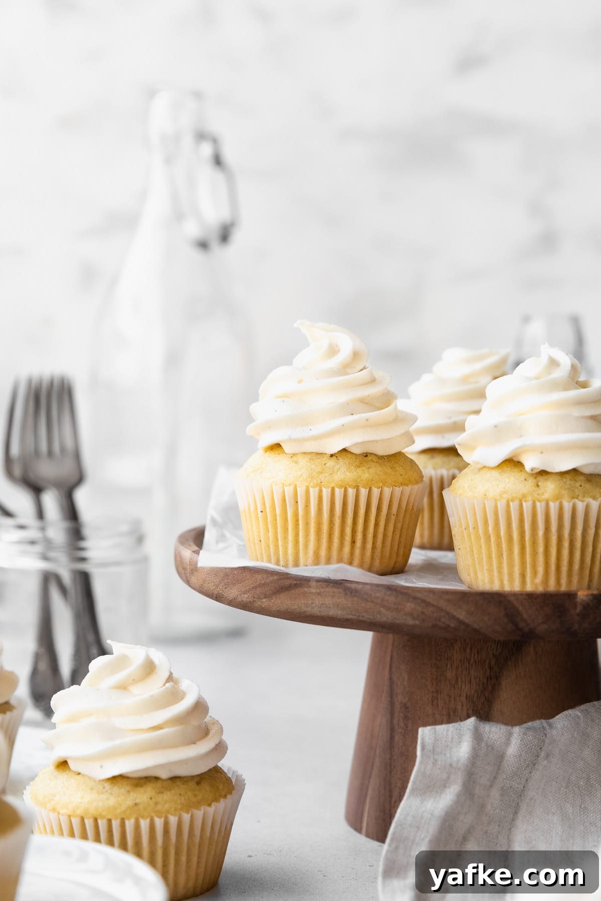 How to Make Dairy-Free Cupcakes 2 Side on shot of dairy-free vanilla cupcakes on a wooden cake stand with a jar of forks and a large jug in the background.
