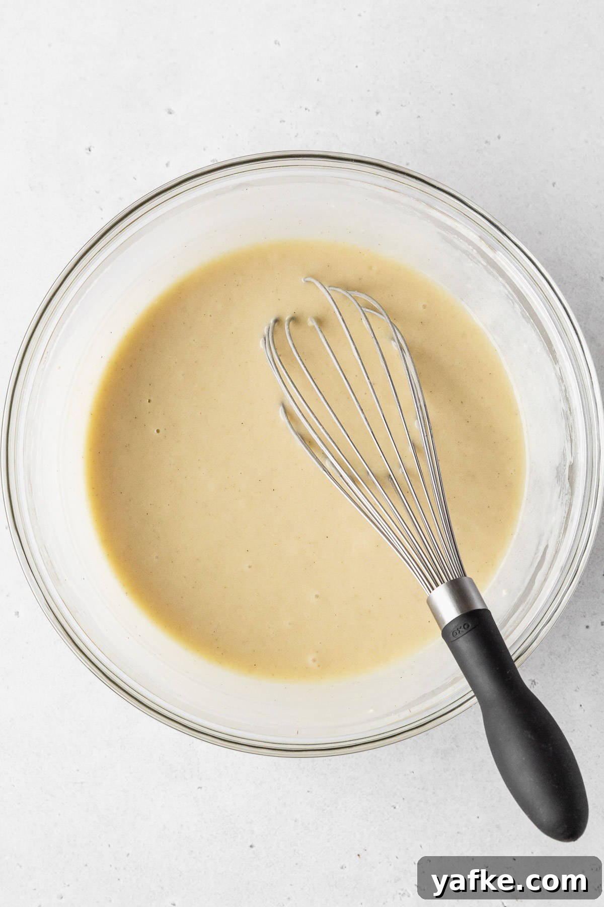 How to Make Dairy-Free Cupcakes 7 Completed dairy free vanilla cupcake batter in a clear mixing bowl with a black-handled whisk.