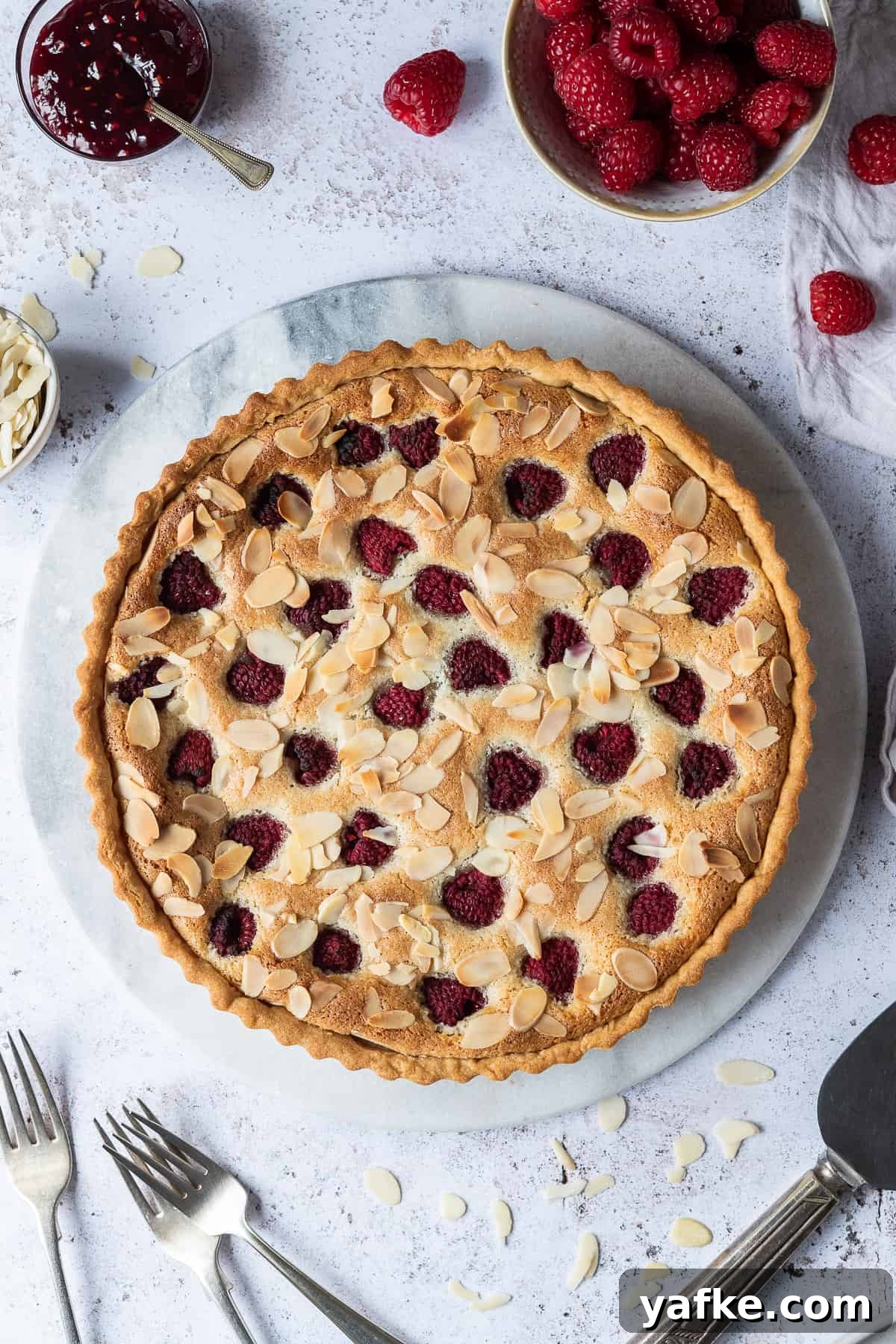 Vegan bakewell tart - a crisp pastry shell filled with raspberry jam, vegan frangipane, fresh raspberries and flaked almonds. Vegan Bakewell tart on a marble board on a white background.