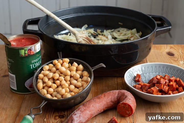 Spanish-Style Chicken, Chorizo, and Chickpea Stew 7 black pan with fried onions a sieve of drained chickpeas 1/2 a chorizo sausages and a can of chopped tomatoes sitting on a wooden kitchen bench