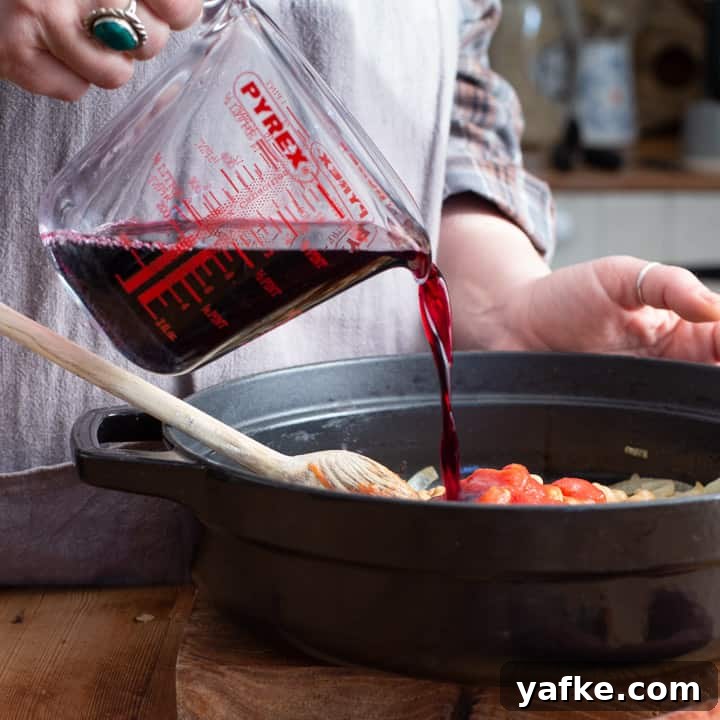 Spanish-Style Chicken, Chorizo, and Chickpea Stew 8 womans hands pouring red wine into a black cast iron casserole dish of chicken chorizo stew