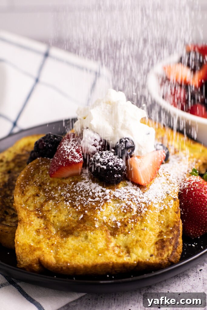 Cinnamon French Toast Recipe with Maple Syrup Glaze 4 Powdered sugar being sprinkled onto 3 pieces of cinnamon French toast on a black plate topped with mixed berries and whipped cream