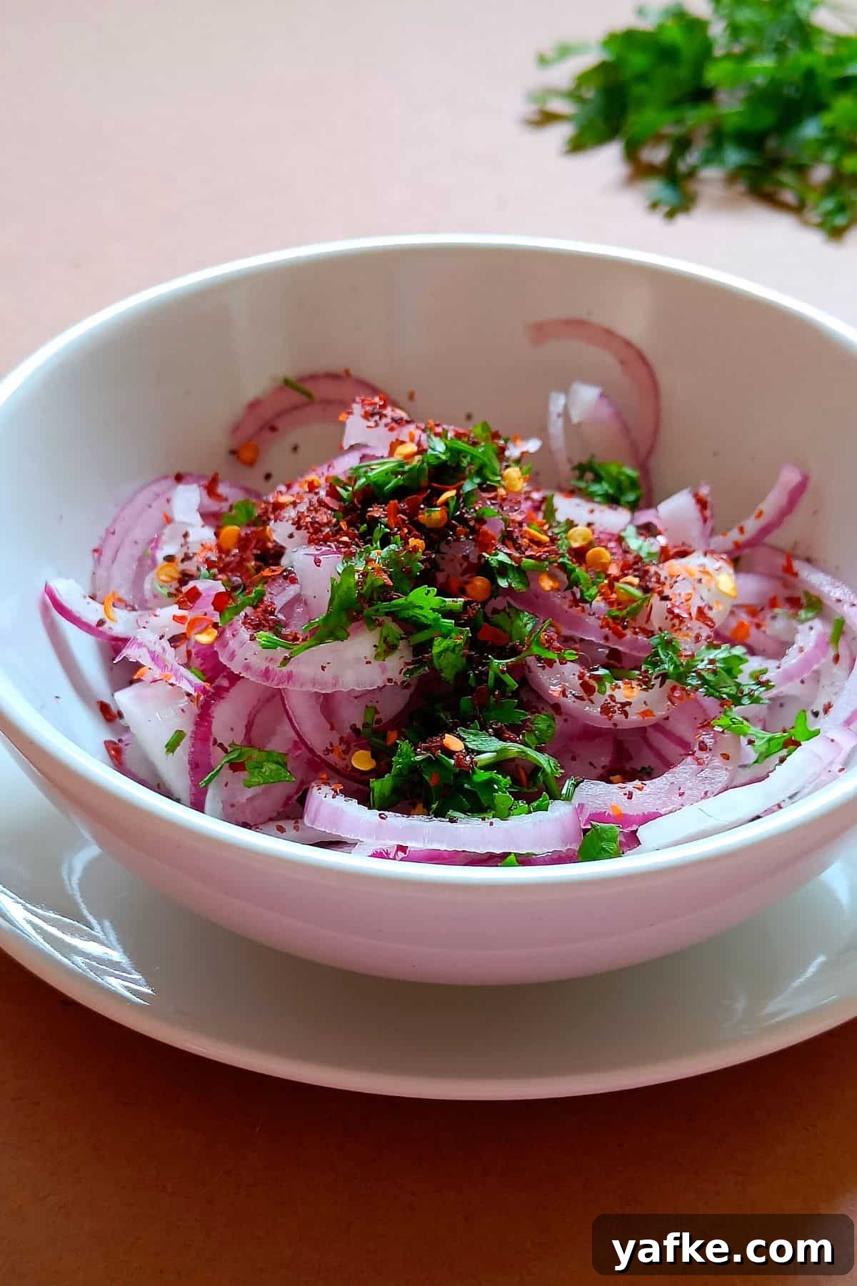Turkish Sumac Marinated Red Onions Recipe 5 Image showing red onions slices, sumac, parsley, chili flakes, and all other ingredients in a white bowl.
