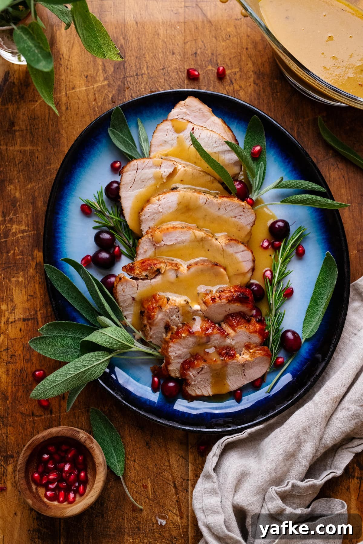 Juicy Maple Herb Air Fryer Turkey Breast 2 A blue plate with slices of air fryer turkey breast and gravy on it with fresh herbs arranged around it. The plate of turkey is arranged on a wooden background with herbs, gravy, and a gray napkin displayed around it.