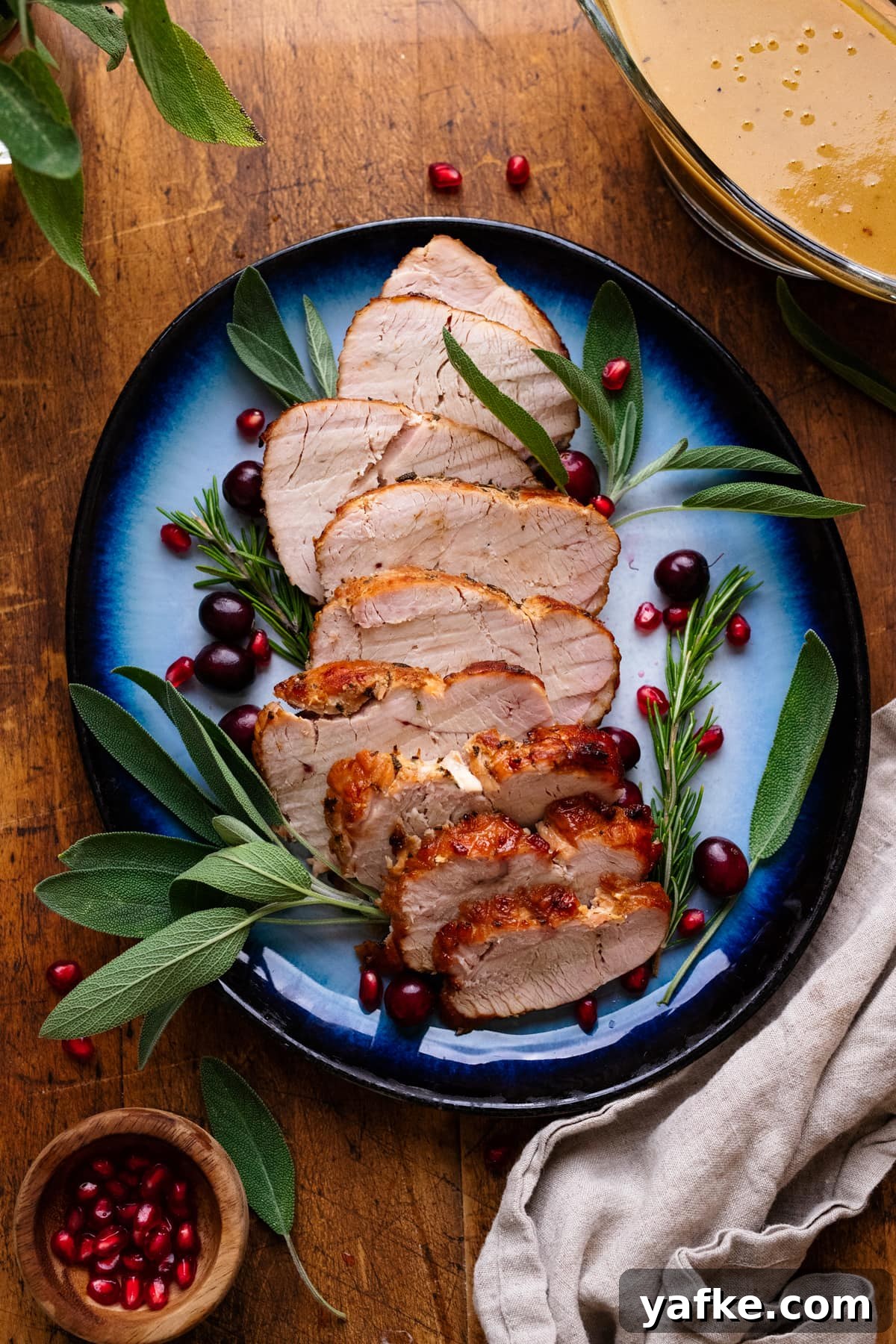 Juicy Maple Herb Air Fryer Turkey Breast 8 A blue plate with slices of air fryer turkey breast on it with fresh herbs arranged around it. The plate of turkey is arranged on a wooden background with herbs, gravy, and a gray napkin displayed around it.