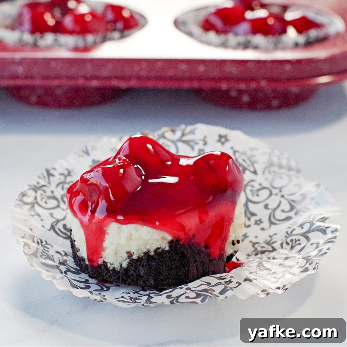 21 Patriotic Canada Day Desserts 10 a mini cherry cheesecake on a damask print muffin wrapper, with a red speckled muffin tin ofmini cherry cheesecakes in the background