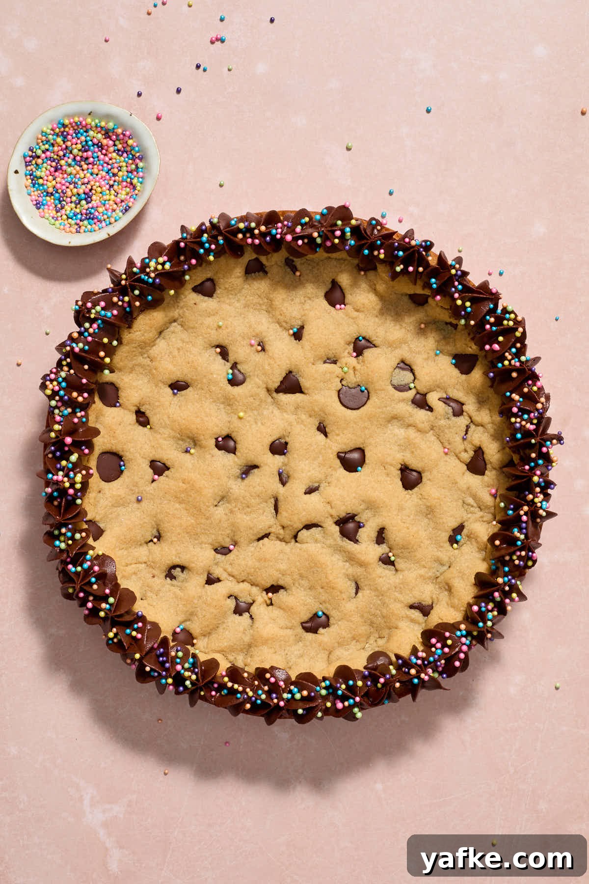 Almond Flour Cookie Cake with Peanut Butter and Chocolate Chips 13 Frosting piped around the edges of the cookie cake with sprinkles on the side.