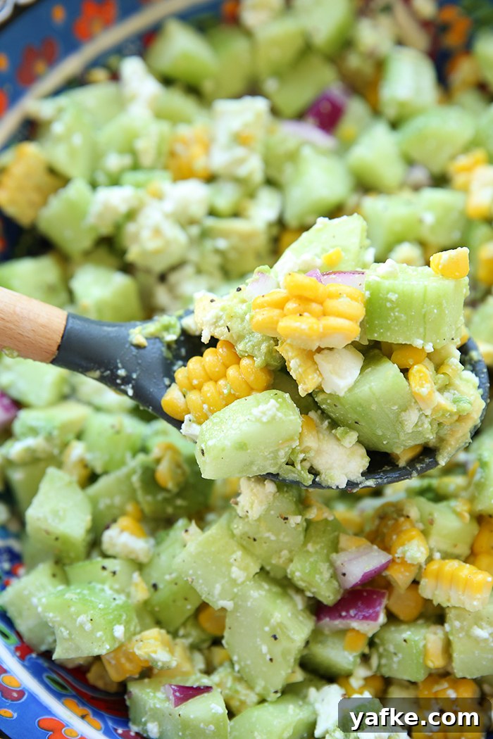 A large bowl of summer cucumber salad with feta and lemon with a spoon held above from the left with salad on it.