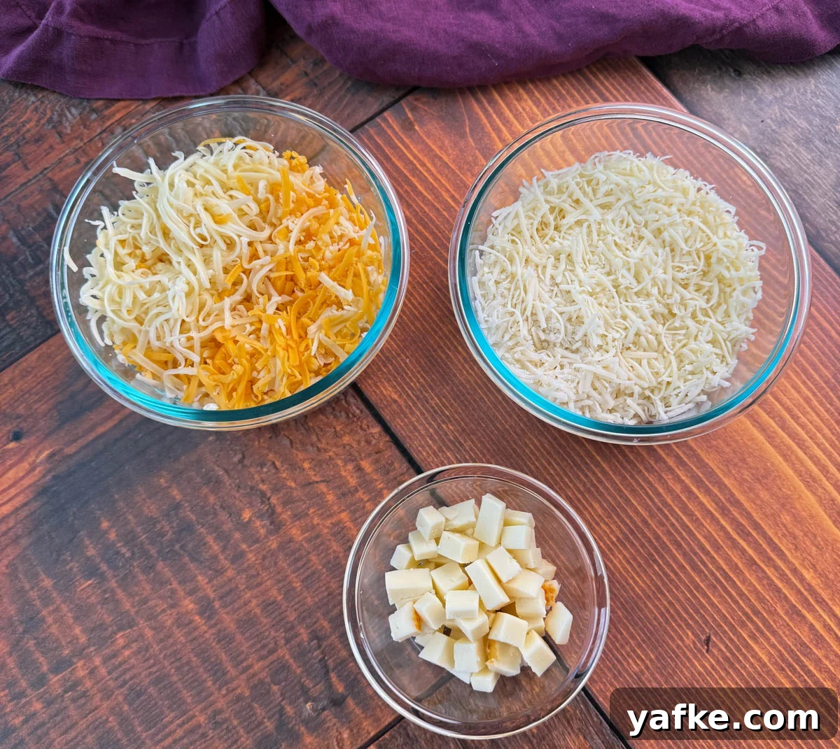 Southern Soul Food Baked Mac and Cheese Recipe 6 Overhead view of three glass bowls on a wooden table—one with shredded cheddar and mozzarella, one with shredded white cheese, and one small bowl with cubed cheese.