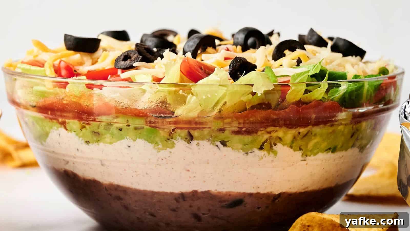 21 Fall Snacks I Tried: 9 Keepers 15 A clear glass bowl with 7 Layer Dip featuring beans, white cream, guacamole, diced tomatoes, shredded cheese, lettuce, and black olives.