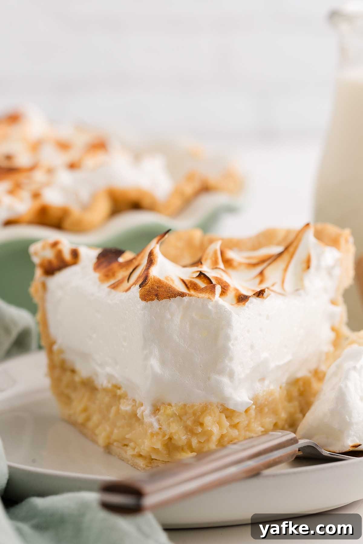 Coconut Meringue Pie with Toasted Coconut Topping 11 A slice of coconut meringue pie with toasted meringue topping sits on a plate, a fork beside it.