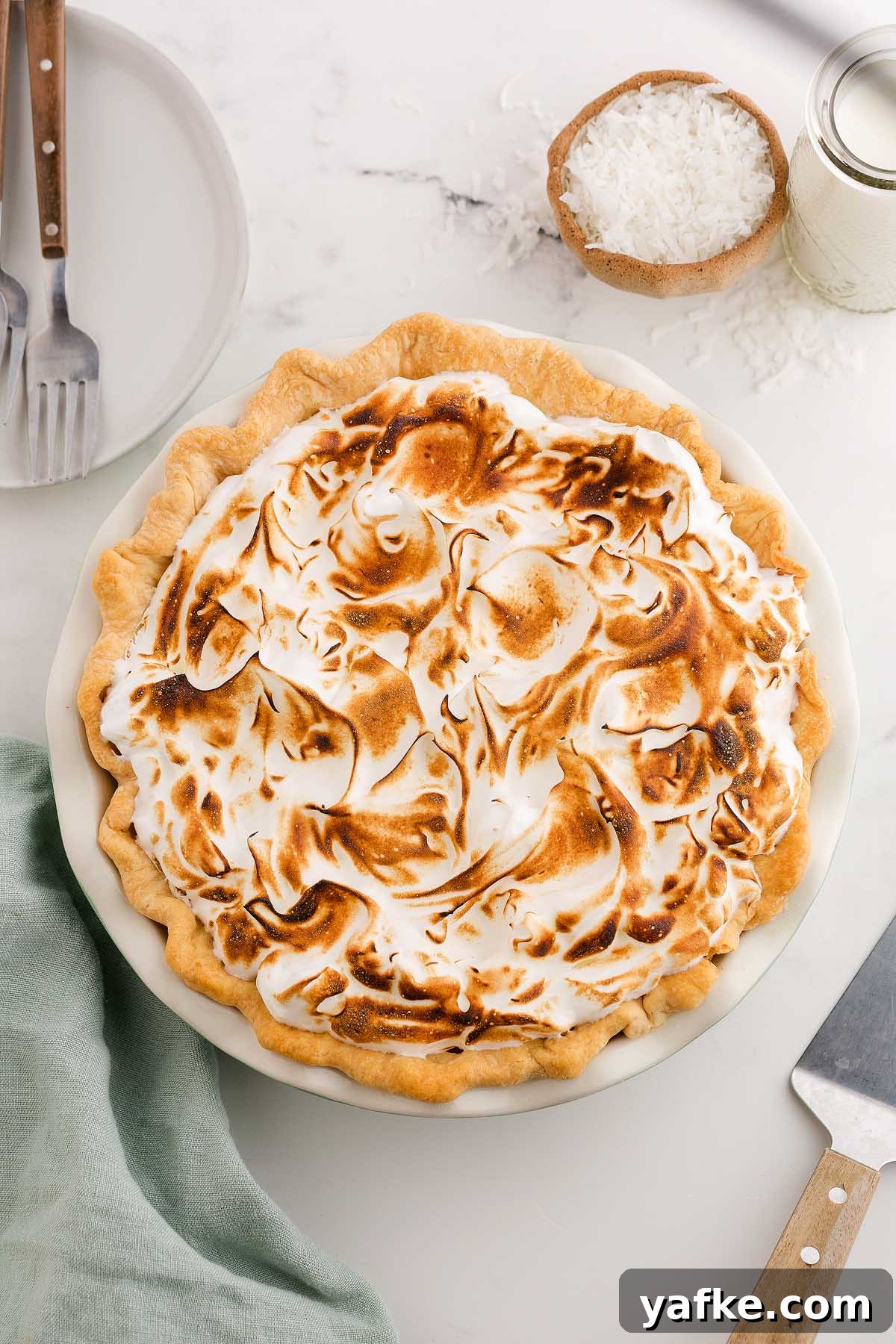 Coconut Meringue Pie with Toasted Coconut Topping 12 A toasted coconut meringue pie with swirled meringue on top, set on a white table alongside plates and utensils.