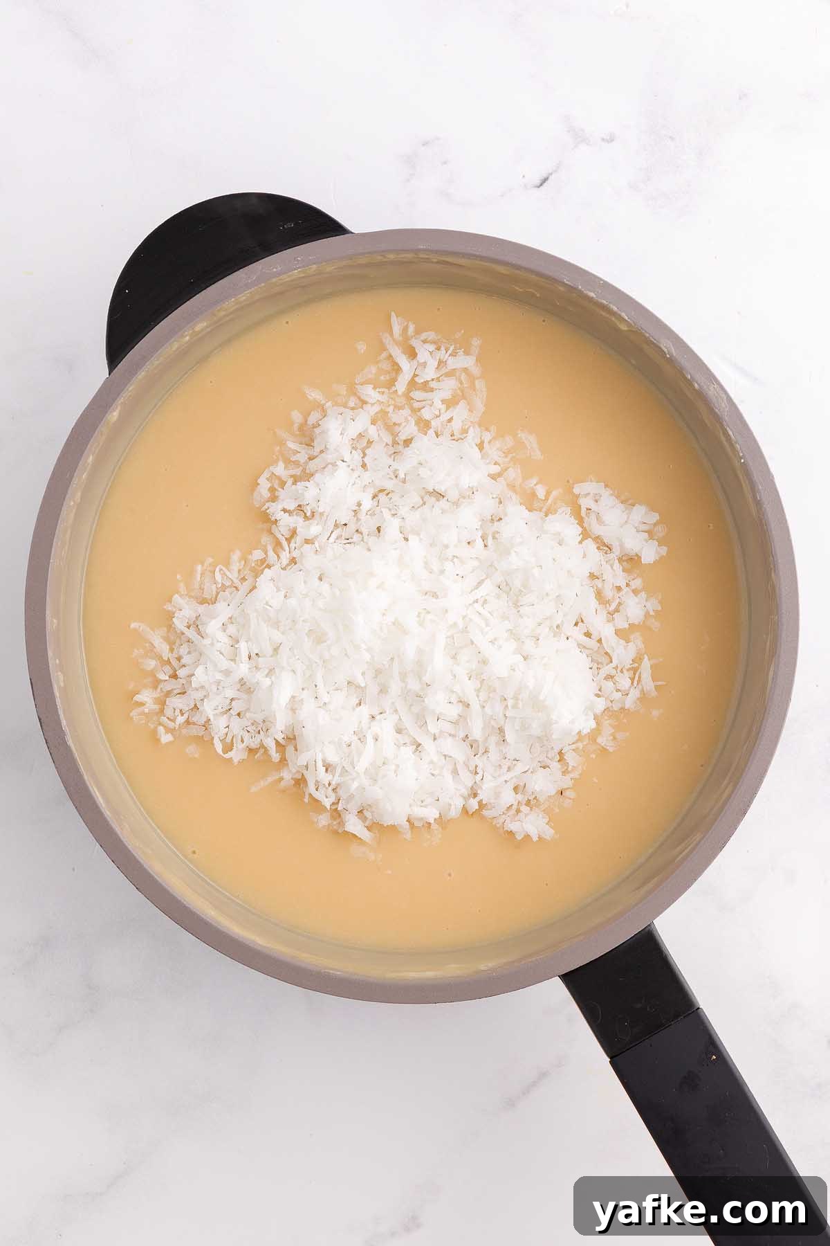 Coconut Meringue Pie with Toasted Coconut Topping 16 A saucepan filled with a creamy coconut meringue pie mixture, topped with shredded coconut, on a white surface.