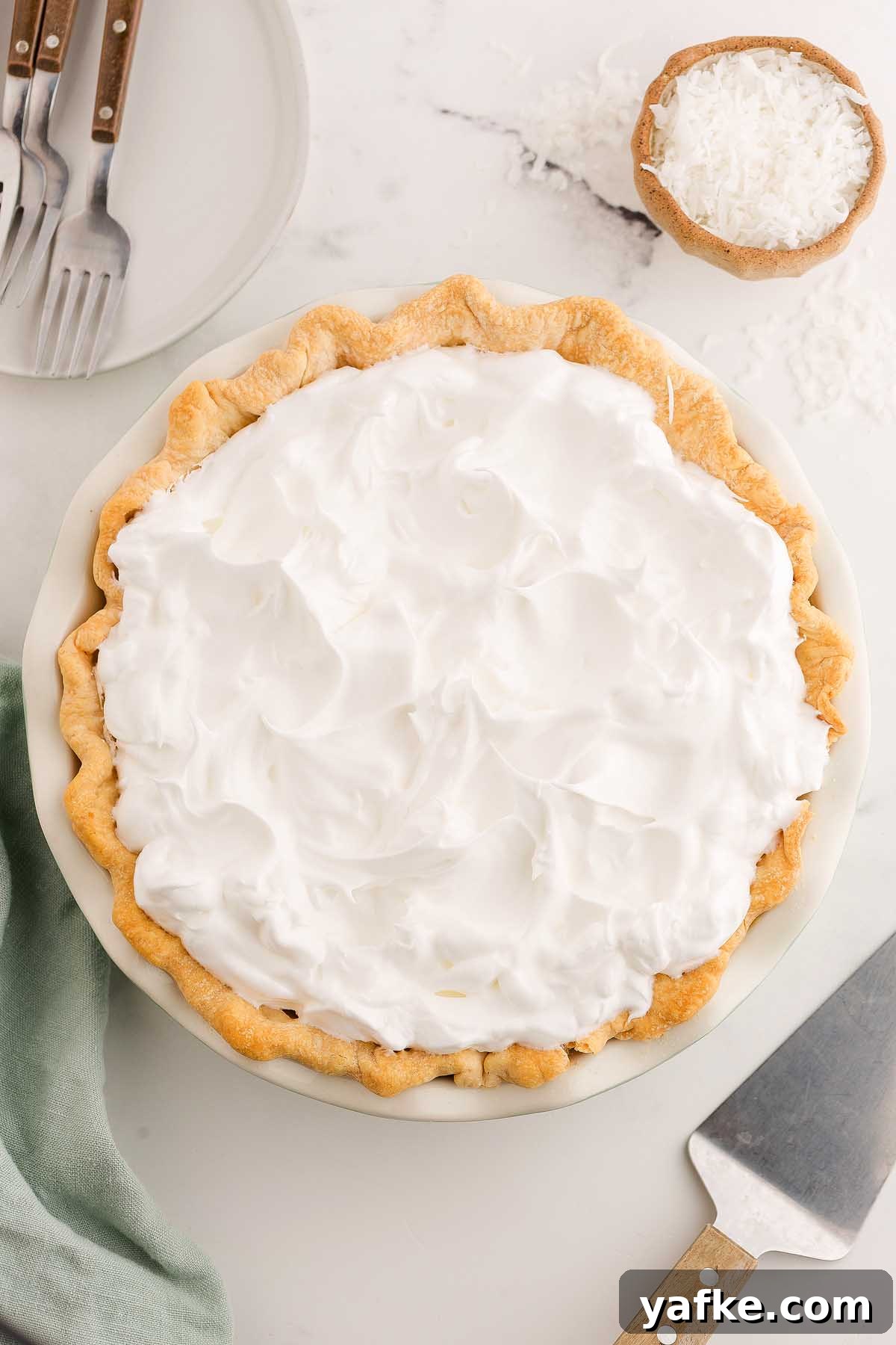 Coconut Meringue Pie with Toasted Coconut Topping 17 A coconut meringue pie topped with fluffy whipped cream sits on a marble surface, with plates and shredded coconut nearby.