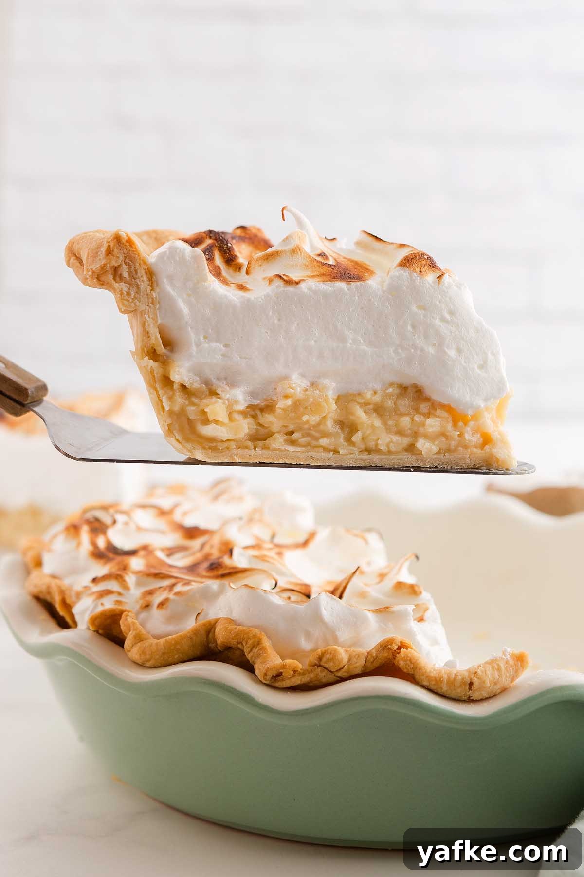 Coconut Meringue Pie with Toasted Coconut Topping 4 A slice of coconut meringue pie, crowned with golden peaks, is being lifted from a green pie dish.