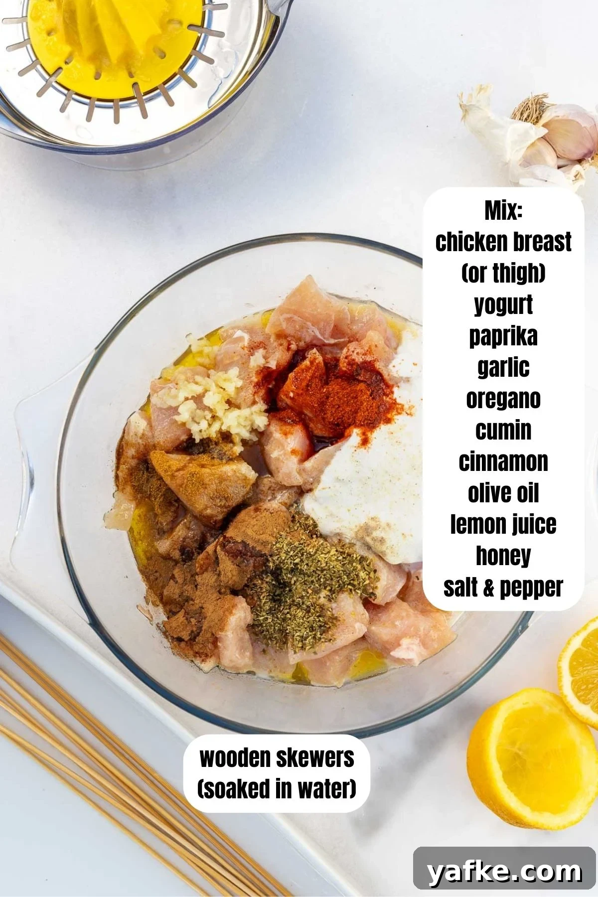 Oven-Baked Greek Chicken Kabobs Recipe 2 Overhead view of the ingredients needed for baked chicken kabobs including a bowl with chicken cubes and all the marinade ingredients, wooden skewers soaking in water and squeezed lemon and garlic around them on a marble background.