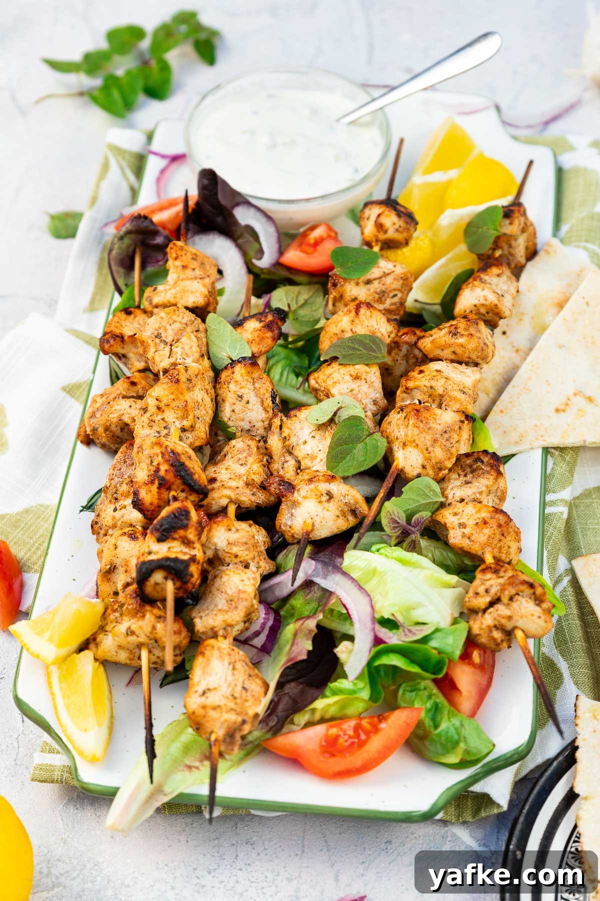 Oven-Baked Greek Chicken Kabobs Recipe 5 A rectangular platter of baked chicken kabobs on a bed of simple salad and with lemon segments and a white dip on a green and white tea towel and on a pale textured background.