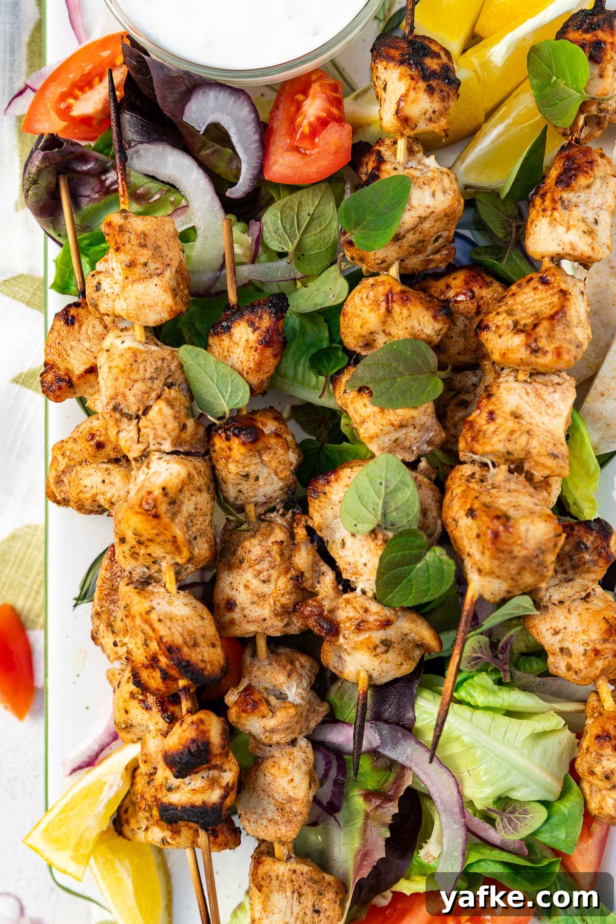 Oven-Baked Greek Chicken Kabobs Recipe 6 Overhead closeup of a rectangular platter of baked chicken kabobs on a bed of simple salad and with lemon segments and a white dip on a green and white tea towel and on a pale textured background.