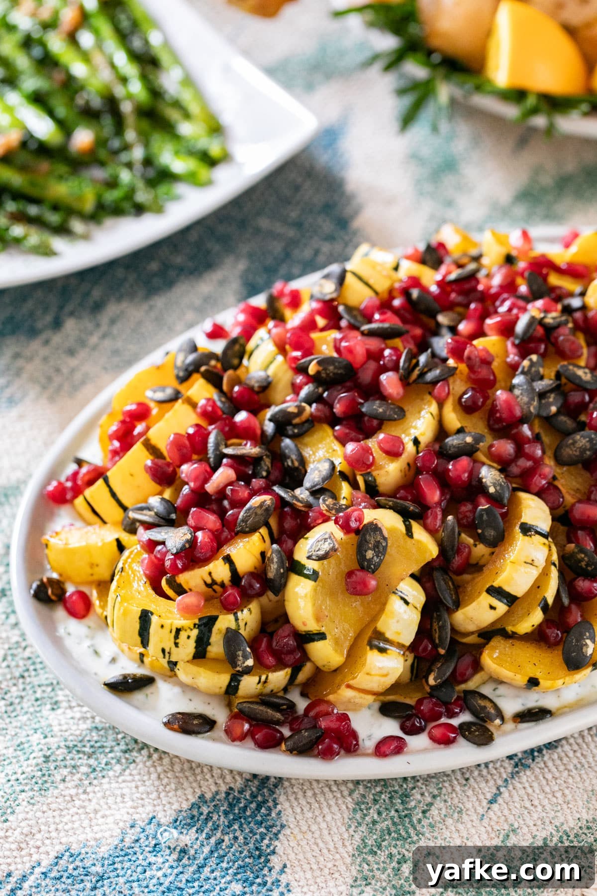 Roasted Delicata Squash with Herbed Yogurt Recipe 11 A plate of squash with yogurt, pomegranate arils and pepitas with a platter of asparagus behind it.