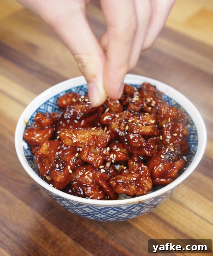 Crispy Sesame Chicken Recipe 8 A hand sprinkles sesame seeds over a finished bowl of saucy sesame chicken.