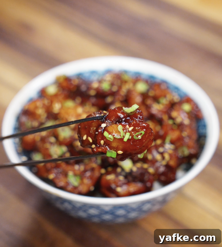 Crispy Sesame Chicken Recipe 2 A close-up of glossy sesame chicken held between chopsticks, showing the sticky sauce, sesame seeds, and scallions.