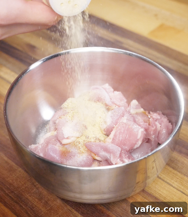 Crispy Sesame Chicken Recipe 3 Raw chicken thigh pieces in a metal bowl being sprinkled with salt, white pepper, garlic powder, and sesame oil.