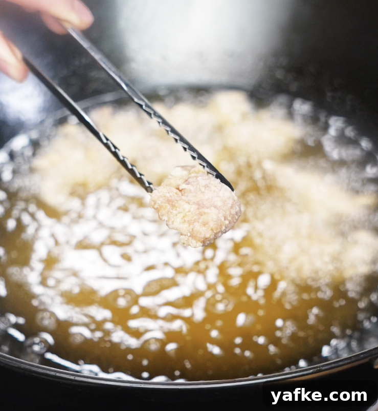 Crispy Sesame Chicken Recipe 5 A piece of lightly fried chicken is lifted from hot oil, showing the pale first-fry texture before crisping.