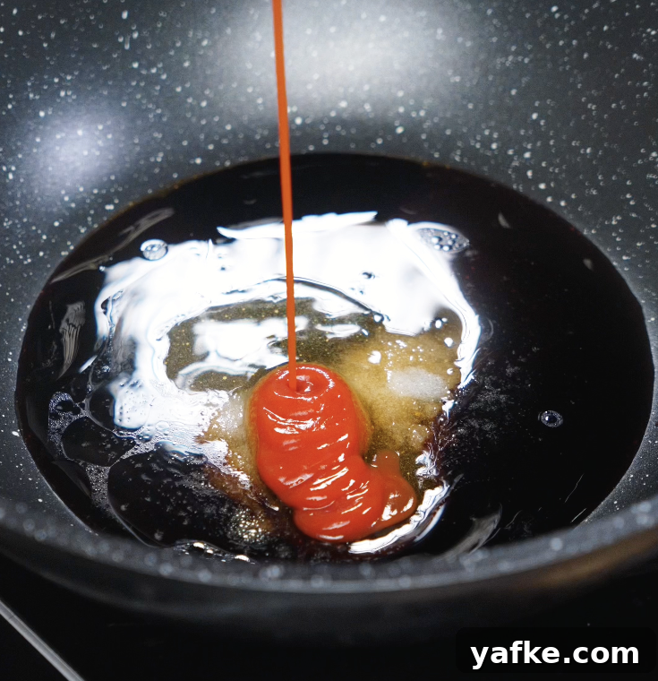 Crispy Sesame Chicken Recipe 7 Thick ketchup being poured into a mixture of soy sauce, vinegar, sugar, and sesame oil in a nonstick pan.