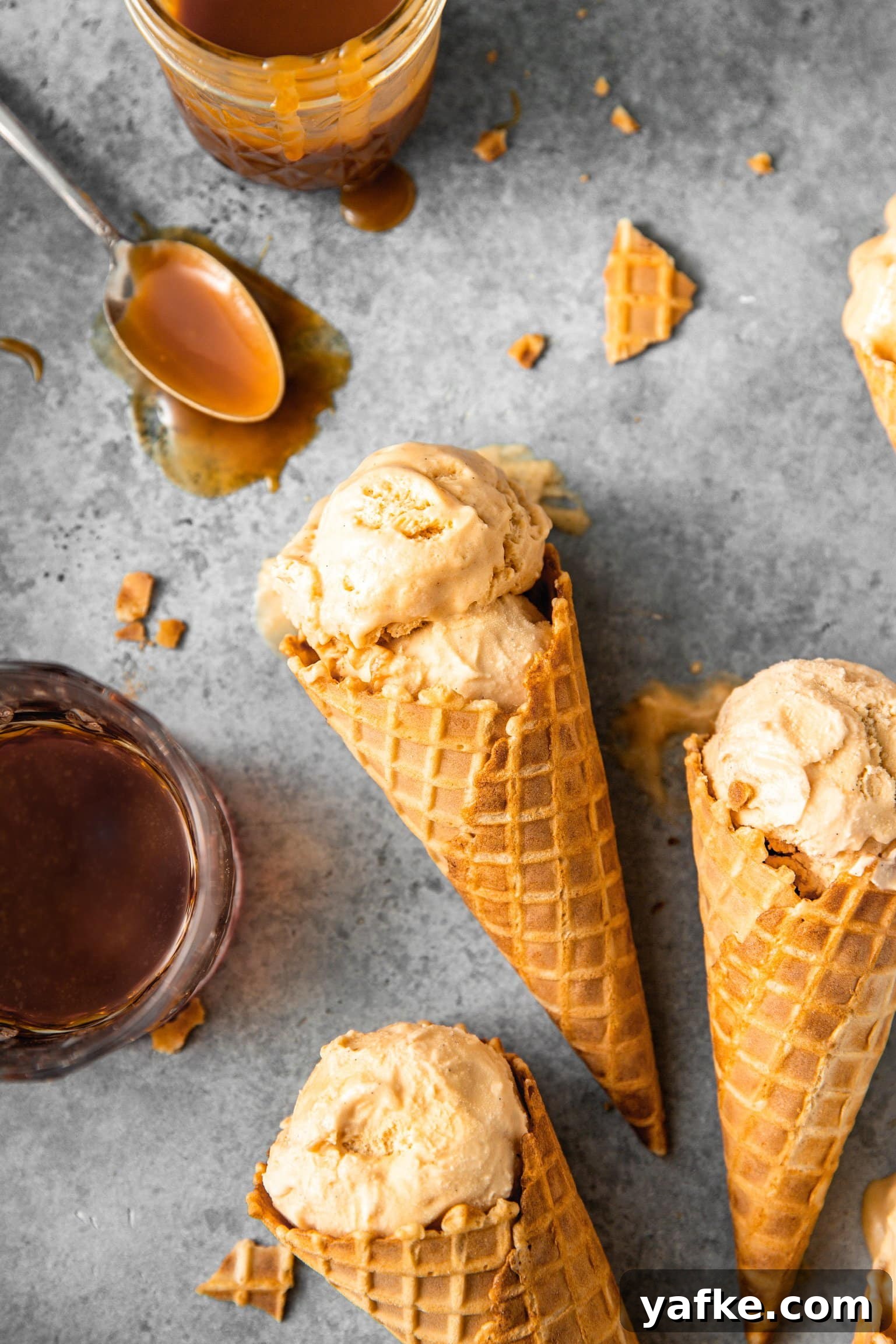 How to Make No Churn Bourbon Salted Caramel Ice Cream 6 Close up of a double scoop of bourbon salted caramel ice cream inside of an ice cream cone with a glass of bourbon and mason jar of bourbon caramel nearby.