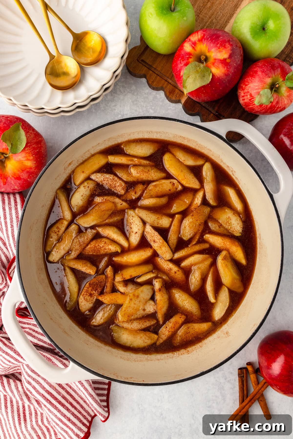 Skillet Fried Apples with Cinnamon and Brown Sugar 5 Fried apples simmering in cinnamon sauce inside a white pot, surrounded by apples and cinnamon sticks.