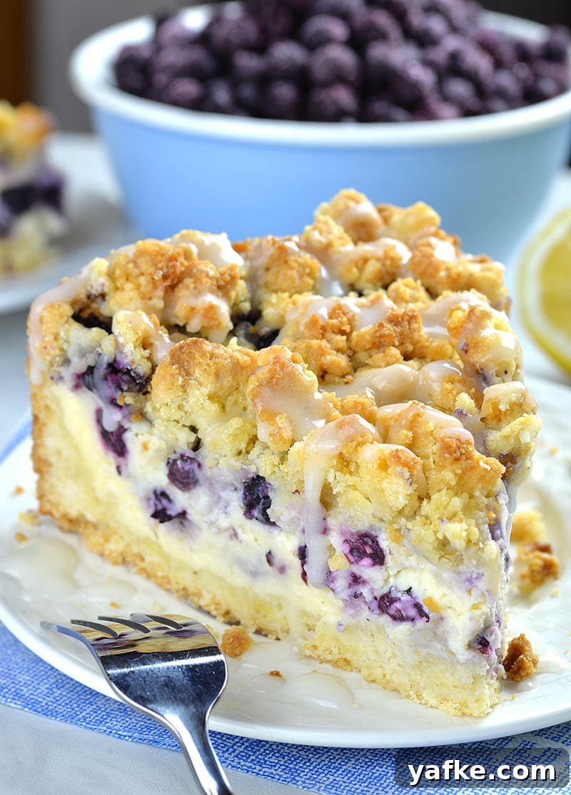 Piece of Blueberry Cheesecake Crumb Cake on a white plate in front of bowl full of fresh blueberries.