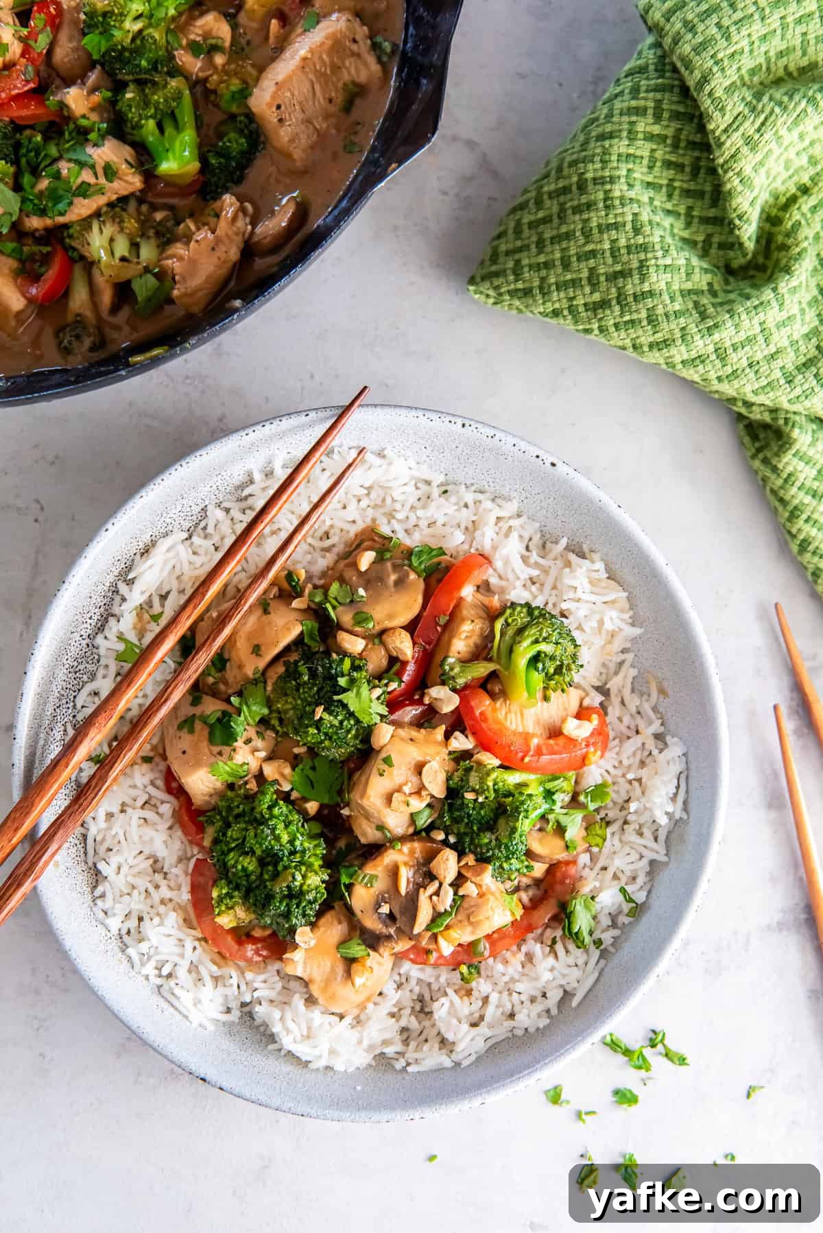 Peanut Chicken Stir-Fry Recipe with Vegetables 2 Chopsticks resting on a bowl of rice topped with peanut chicken stir fry next to a skillet and green kitchen cloth.
