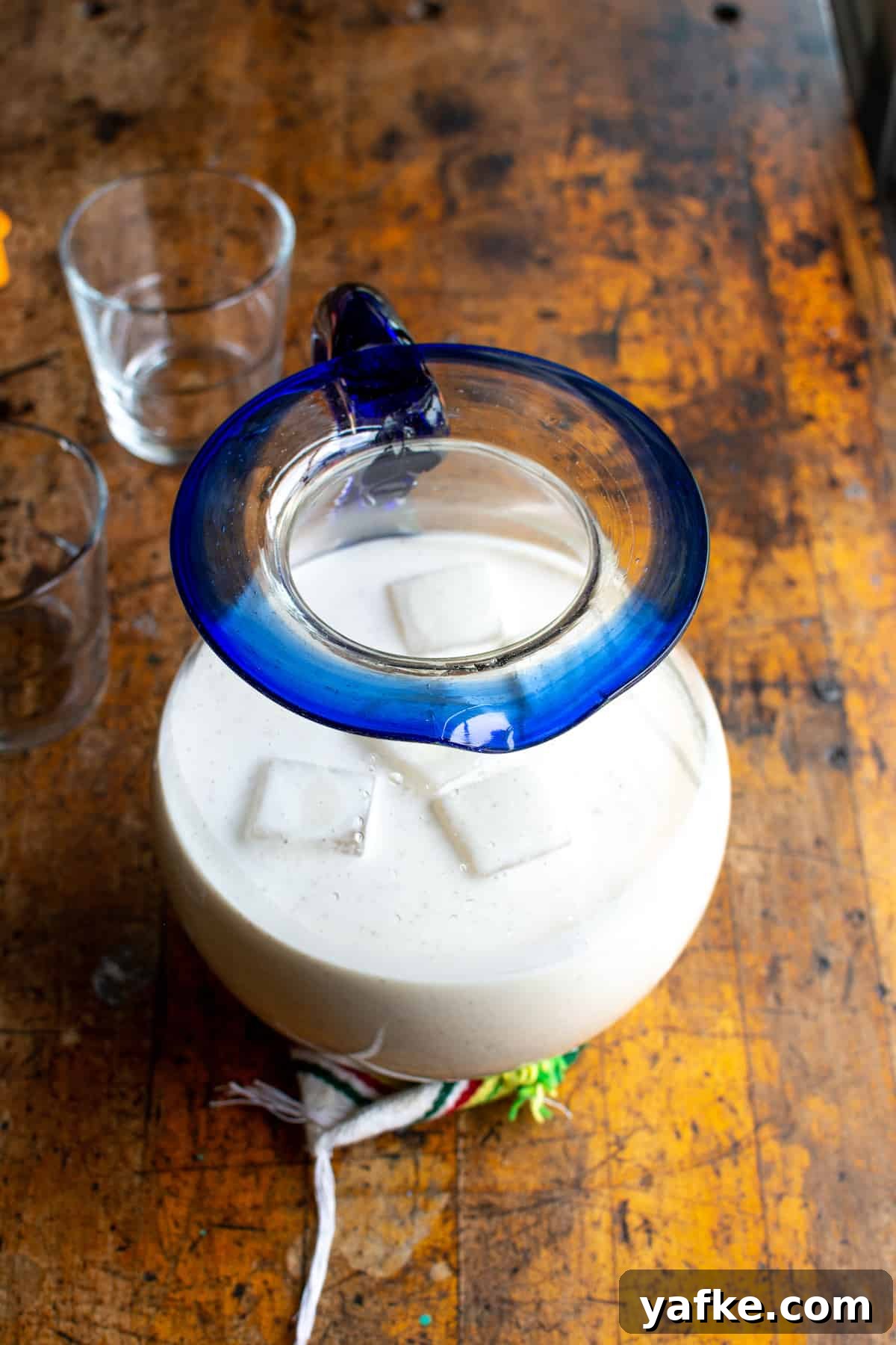 Authentic Creamy Mexican Horchata Recipe with Rice and Cinnamon 3 A glass pitcher with a blue rim filled with horchata and ice cubes sitting on a wood table with a couple of glasses next to it.