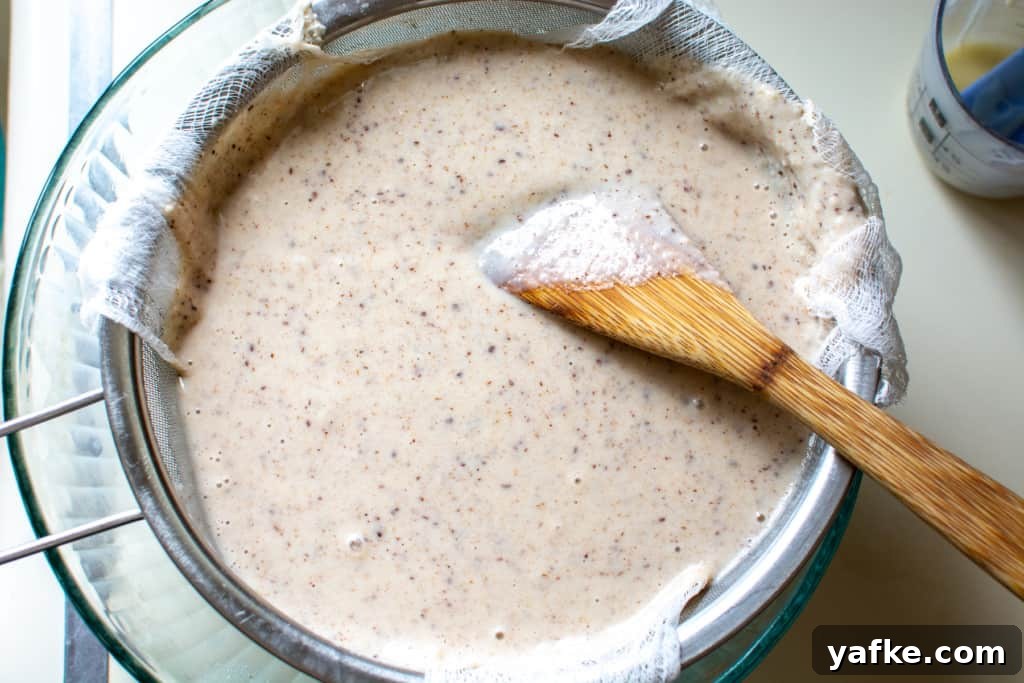 Authentic Creamy Mexican Horchata Recipe with Rice and Cinnamon 8 A glass bowl with a strainer lined with cheesecloth over the bowl and a wood spoon in the bowl. The strainer is filled with horchata.