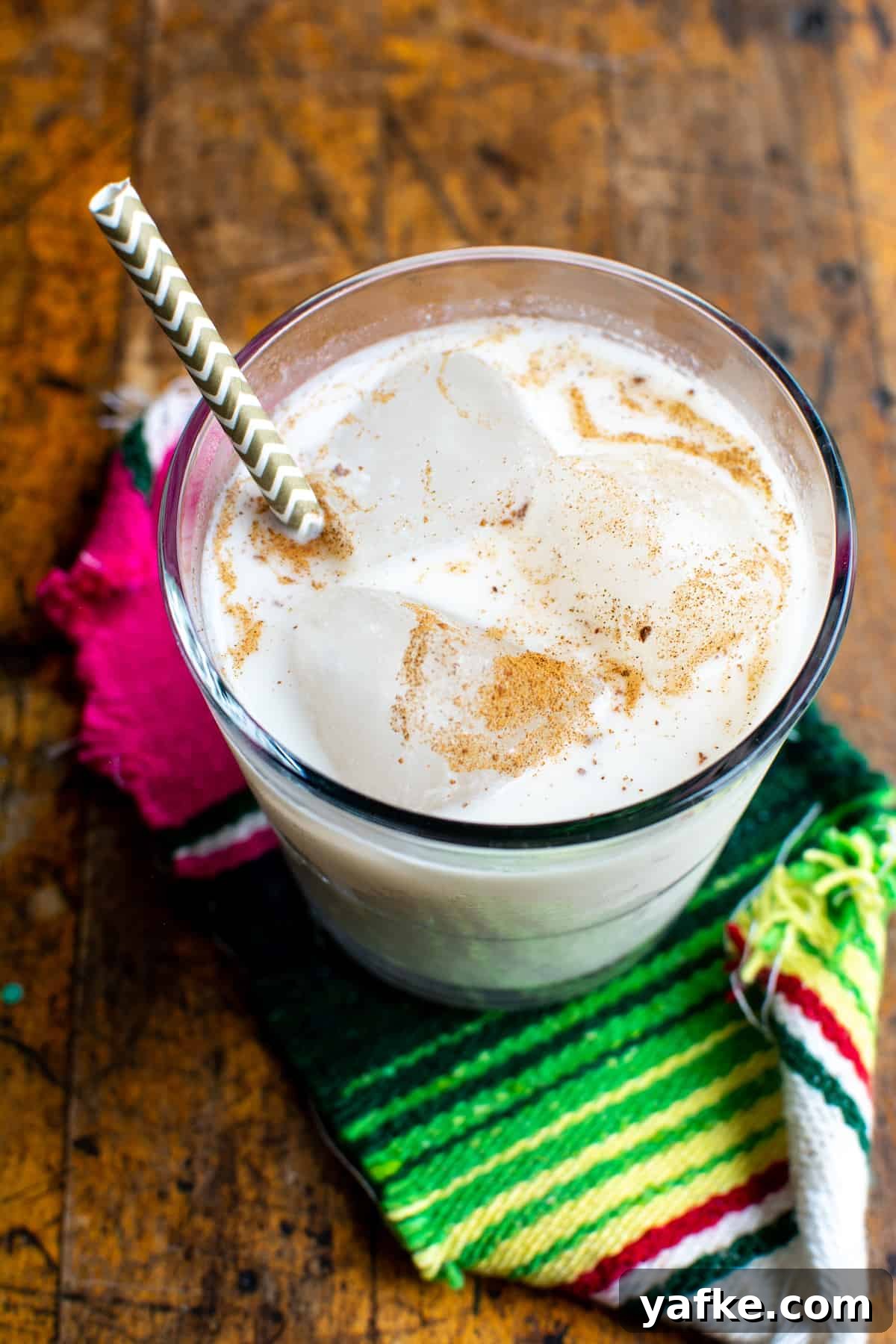 Authentic Creamy Mexican Horchata Recipe with Rice and Cinnamon 9 A glass filled with horchata and ice and a gold striped straw. The top is sprinkled with cinnamon and sitting on a wood table.