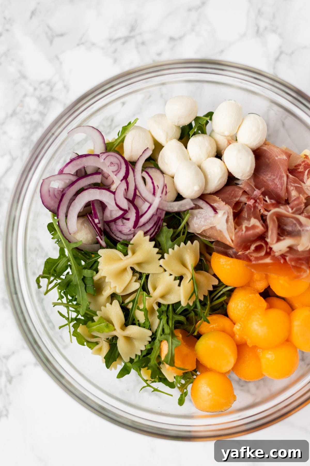 Prosciutto melon pasta salad ingredients in a glass bowl.