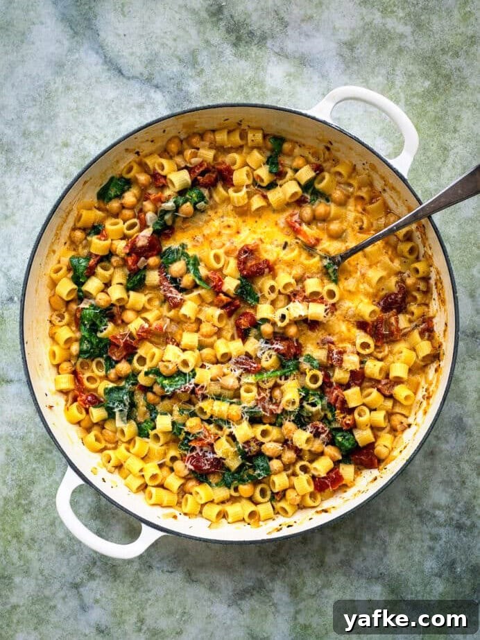 Marry Me Chickpeas One-Pot Pasta Recipe 6 A large white pan filled with creamy pasta, sun-dried tomatoes, spinach, and Marry Me Chickpeas, topped with grated cheese, sits on a textured green surface. A metal spoon rests inside the pan.