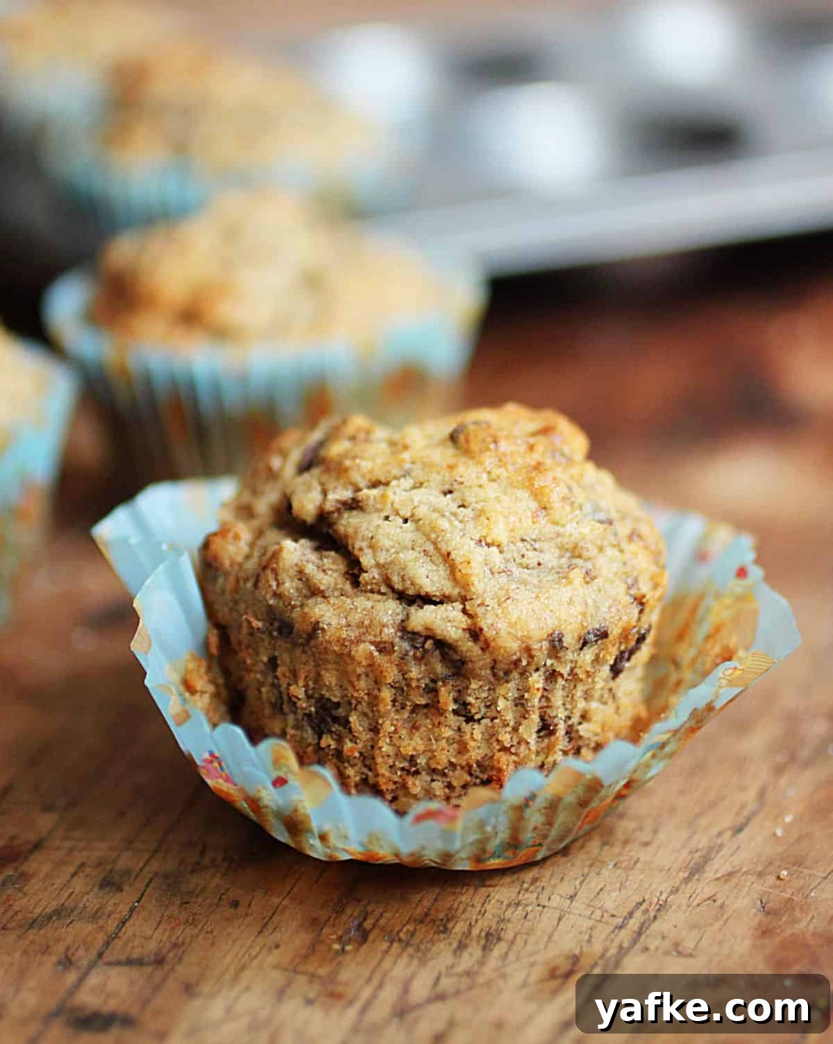 Gluten-Free Banana Chocolate Chip Muffins Recipe 2 Whole banana muffin in opened paper liner on wooden table. More muffins and pan in background.