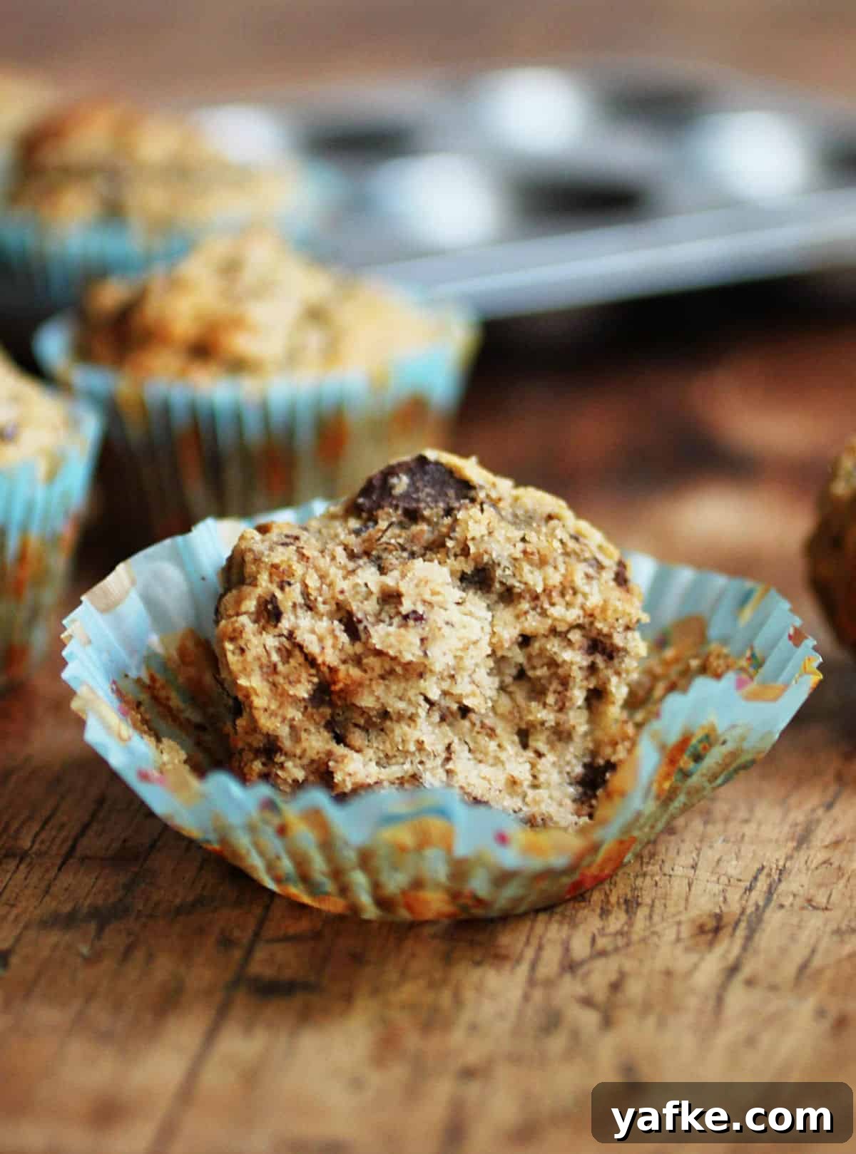 Banana Chocolate Pecan Quinoa Muffins - Gluten Free Half a banana chocolate chip muffin in opened blue paper liner on a wooden board. Pan and muffins in background.