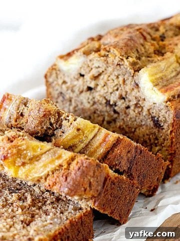 White parchment paper with loaf of banana bread with some cut slices, white background.