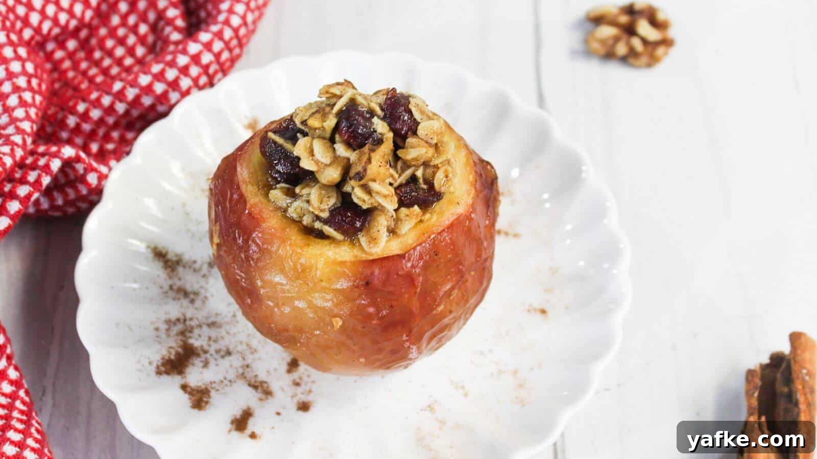 11 Air Fryer Dessert Recipes That Will Make You Ditch the Oven 6 Baked apple on a white plate filled with oats, nuts, and cranberries, with cinnamon sprinkled around. A red checkered cloth is partially visible on the side.