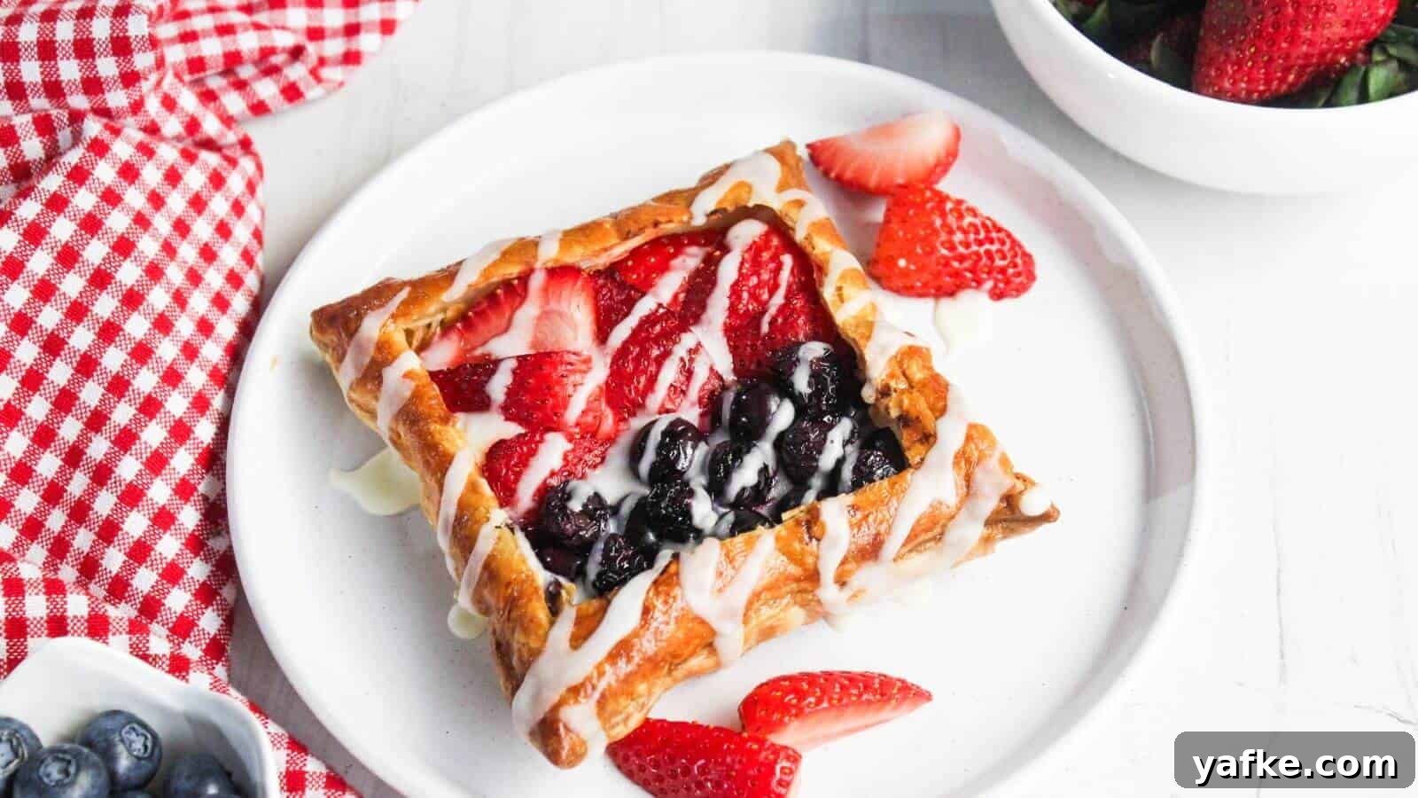 11 Air Fryer Dessert Recipes That Will Make You Ditch the Oven 7 A pastry with berry filling and white drizzle on a white plate is accompanied by strawberry slices. A red and white checkered cloth, blueberries, and a bowl of strawberries are nearby.