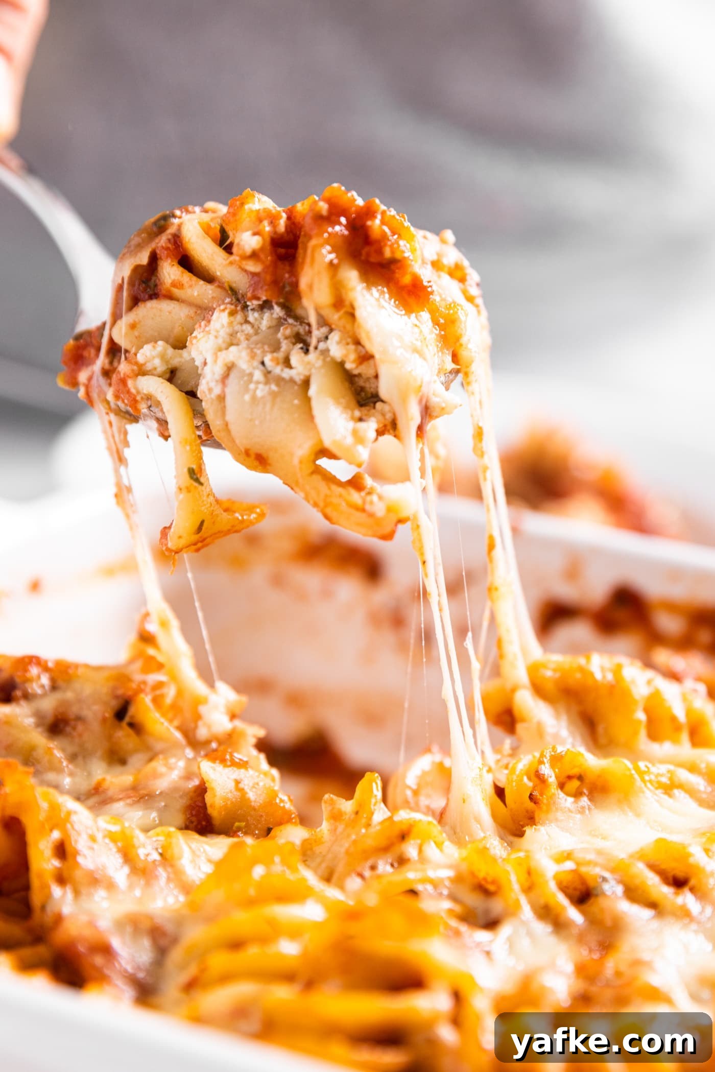 Cheesy Baked Rotini Casserole with Marinara and Mozzarella 2 Spoon pulling piece of baked rotini casserole from baking dish with cheese strings clinging to the rest of the casserole