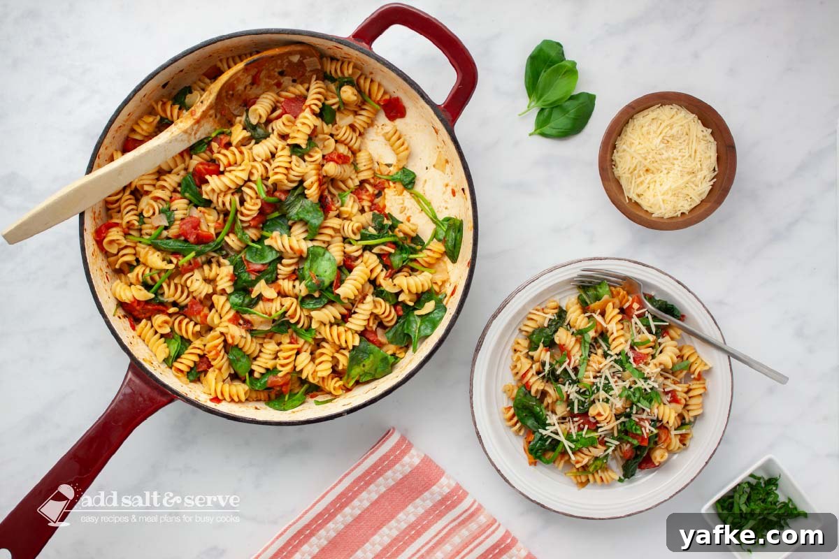 One-Pot Tomato and Spinach Pasta Recipe 2 Plate of tomato Spinach Pasta garnished with Parmesan cheese and fresh basil strips on a counter beside the pan of the full dish.